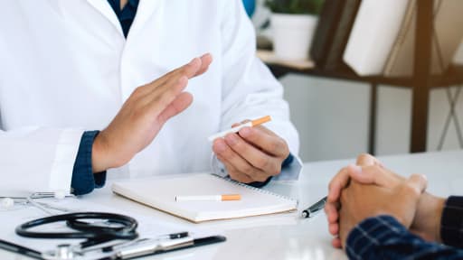 Schreibtisch eines Arztes mit zwei Zigaretten, während er einem Patienten gegenübersitzt und im Gespräch gestikuliert.