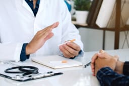 Schreibtisch eines Arztes mit zwei Zigaretten, während er einem Patienten gegenübersitzt und im Gespräch gestikuliert.